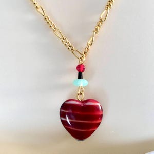 Puede incluir: Un collar de cadena dorada con un colgante de corazón a rayas rojas y blancas. El colgante está acentuado con pequeñas cuentas rojas, negras y turquesas.