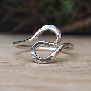 May include: A silver ring with a hammered texture, featuring a flowing, abstract design. The ring's shape curves in a continuous, wave-like pattern. The ring is displayed on a wooden surface.