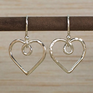 May include: A pair of silver heart-shaped earrings with a twisted wire detail. The earrings are hanging from a wooden surface.