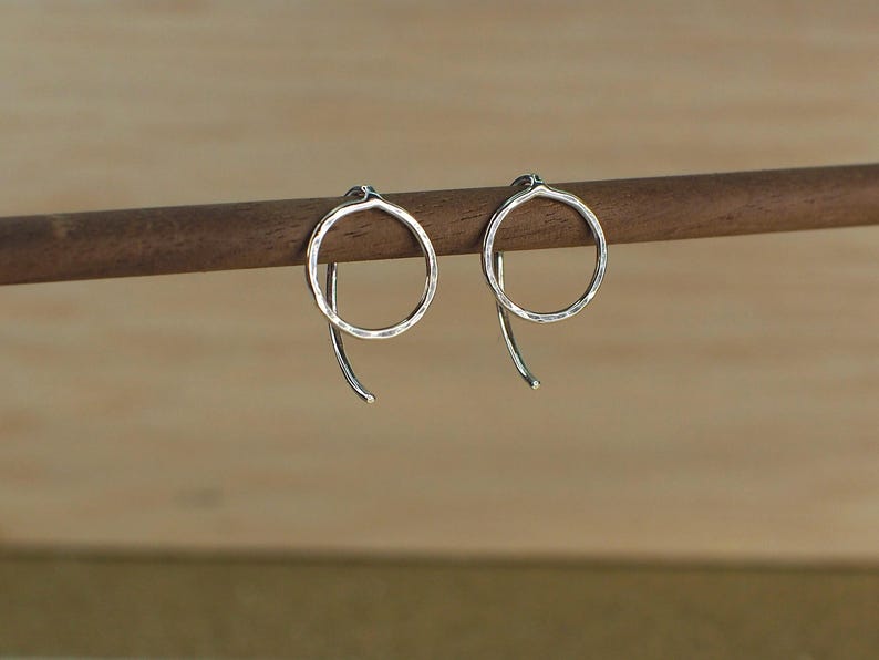Two minimalist sterling silver circle earrings with a hammered textured surface. The earrings are hanging from a wooden surface and catch the light.