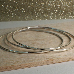 May include: Three silver bangles stacked on a wooden surface. The bangles are smooth and have a slightly hammered texture.