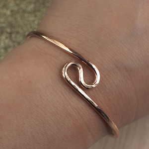 May include: A copper-coloured cuff bracelet with a hammered texture. The open design features a swirling, decorative element. The bracelet is worn on a wrist, showcasing its simple yet elegant style.