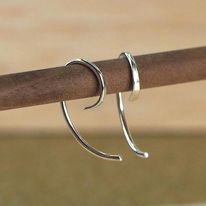 May include: A pair of silver-coloured, curved earrings. Each earring has a hook shape that curves downwards, with a smooth, polished finish. The earrings are displayed on a brown wooden dowel, showcasing their minimalist design and reflective surface.