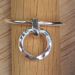 May include: A silver ring featuring a hammered circular pendant. The ring is presented on a wooden dowel, highlighting its design. The ring's band is smooth and connects to the pendant with a small, vertical bar.