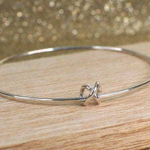 May include: A silver bangle bracelet with a knot design. The bracelet is thin and circular, with a small knot detail at the center. The background is a light brown wooden surface with a gold glittery background.
