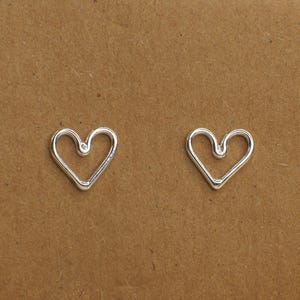 May include: Two silver heart-shaped charms. The charms are open, with a simple, elegant design. The hearts are placed on a brown background. These charms could be used for jewelry making or other craft projects.