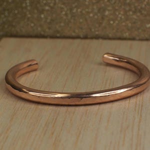 Könnte beinhalten: Ein Kupfer-Armreif mit schlichtem, offenem Design. Das Armband hat eine warme, rötlich-braune Farbe und scheint aus massivem Metall zu bestehen. Es liegt auf einer hellen Holzoberfläche.