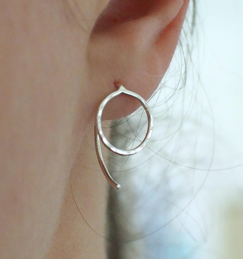 A sterling silver circle earring with a curved hook back. The hoop shaped earring has a polished hammered surface to catch the light. Shown in an earlobe.