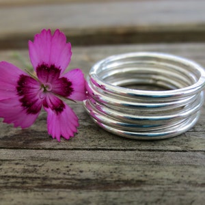 Könnte beinhalten: Ein Stapel aus sechs silbernen Ringen, jeder mit einer leicht unterschiedlichen Breite, in einem Kreis auf einer Holzoberfläche angeordnet. Eine rosa Blume mit einer dunklen Mitte liegt neben den Ringen.