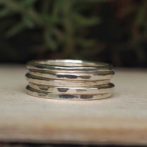 May include: A stack of four silver rings, each featuring a distinct hammered texture. The bands are circular and metallic, reflecting light across their uneven, dimpled surfaces. The rings are positioned one on top of the other in a vertical column.