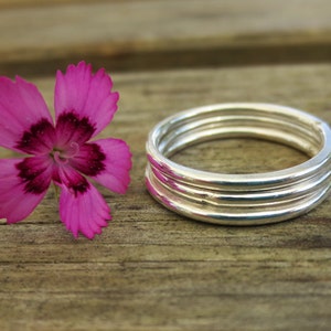 May include: Four silver stacking rings on a wooden surface next to a pink flower.