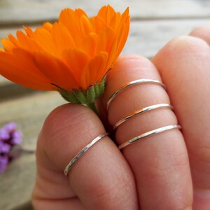 May include: A hand wearing four silver hammered stacking rings. The rings are thin and delicate, and they are stacked on the middle finger. The hand is holding a bright orange flower.