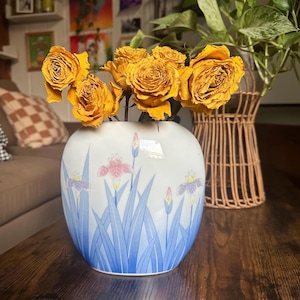 Può includere: Un vaso in ceramica bianca di forma arrotondata con un disegno dipinto di foglie blu e fiori di iris rosa e viola. Il vaso è riempito con diverse rose gialle essiccate che presentano petali color oro intenso e steli visibili.