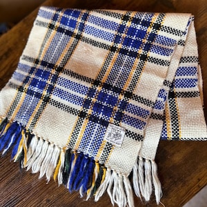 May include: A woven scarf featuring a plaid pattern in cream, royal blue, black, and yellow. The fabric has a visible grid texture and a fringed edge. A small white rectangular label is sewn onto the corner with the text HAND LOOM WOVEN PURE ZEPHYR WOOL.