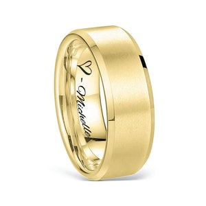 May include: A gold-colored ring with a flat top and beveled edges. The inside of the ring is engraved with a heart symbol and the name "- Michelle". The ring has a smooth, matte finish.