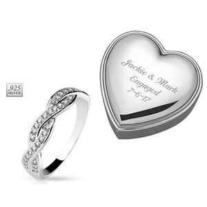 May include: A silver ring with a braided design and small clear stones. The ring is in a heart-shaped box with the inscription "Jackie & Mark Engaged 7-6-17".