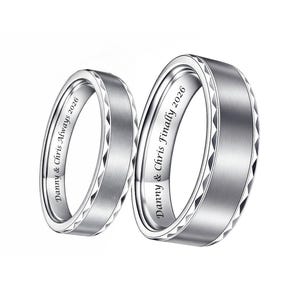 May include: Two silver wedding bands with engraved text. One ring reads "Danny & Chris Always 2026" and the other reads "Danny & Chris Finally 2026". The rings have a brushed metal finish with a geometric design around the edges.