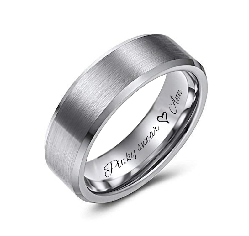 May include: A silver-colored ring with a brushed metal finish. The ring has a flat top and beveled edges. The inside of the ring is engraved with the words "Pinky swear" and "Ann" with a heart symbol. A close-up shot against a white background.