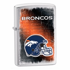 Puede incluir: Un encendedor de metal plateado con el logotipo de los Denver Broncos. El dise&ntilde;o incluye un casco azul marino con una cabeza de caballo naranja, sobre un fondo naranja y negro desgastado. La palabra "BRONCOS" est&aacute; impresa en la parte superior.