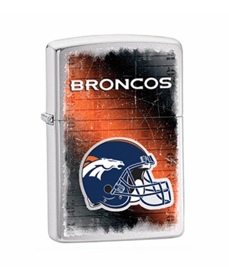 Puede incluir: Un encendedor de metal plateado con el logotipo de los Denver Broncos. El dise&ntilde;o incluye un casco azul marino con una cabeza de caballo naranja, sobre un fondo naranja y negro desgastado. La palabra "BRONCOS" est&aacute; impresa en la parte superior.
