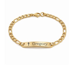 May include: A gold-colored bracelet with a Figaro chain design. The bracelet features a rectangular plate engraved with a cross and the name "Gregory". The bracelet is shown against a white background.
