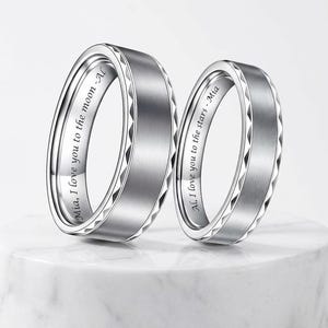 May include: Two silver wedding bands with a brushed metal finish and engraved text. One ring says "Mia, I love you to the moon - Al" and the other says "Al, I love you to the stars - Mia". The rings have a geometric design.