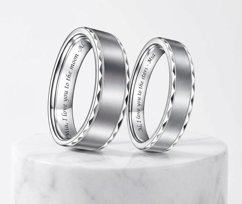 May include: Two silver wedding bands with a brushed metal finish and engraved text. One ring says "Mia, I love you to the moon - Al" and the other says "Al, I love you to the stars - Mia". The rings have a geometric design.