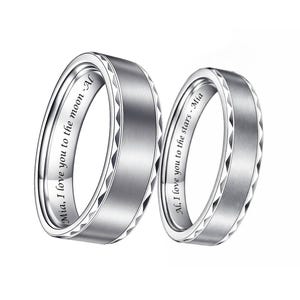 May include: Two silver rings with a brushed metal finish. The rings have a geometric design on the edges and engraved text inside. One ring reads "Mia, I love you to the moon - Al" and the other reads "Al, I love you to the stars - Mia".