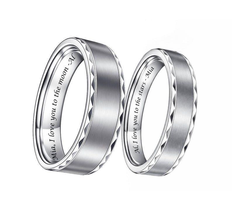 May include: Two silver rings with a brushed metal finish. The rings have a geometric design on the edges and engraved text inside. One ring reads "Mia, I love you to the moon - Al" and the other reads "Al, I love you to the stars - Mia".