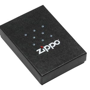 May include: A black textured box with the white "Zippo" logo and a series of small, evenly spaced dots. The Zippo logo is in a clean, sans-serif font, with a red flame detail. The box is rectangular and appears to be made of cardboard.
