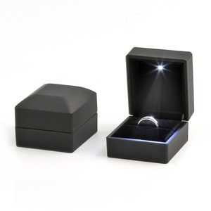 May include: Two black ring boxes, one closed and one open, with a ring illuminated by a spotlight. The open box reveals a silver ring set against a black velvet interior. The closed box is a simple, geometric design.