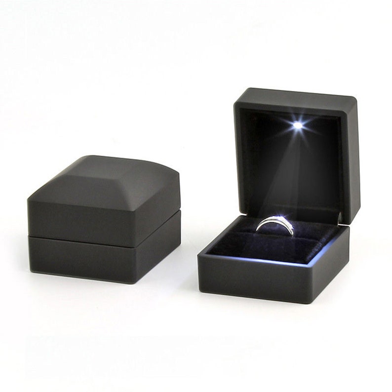 May include: Two black ring boxes, one closed and one open, with a ring illuminated by a spotlight. The open box reveals a silver ring set against a black velvet interior. The closed box is a simple, geometric design.