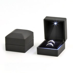 May include: Two silver rings displayed in an open black ring box with a spotlight shining on them. The box is velvet-lined and has a hinged lid. A closed black box is next to the open box. The boxes are on a white background.