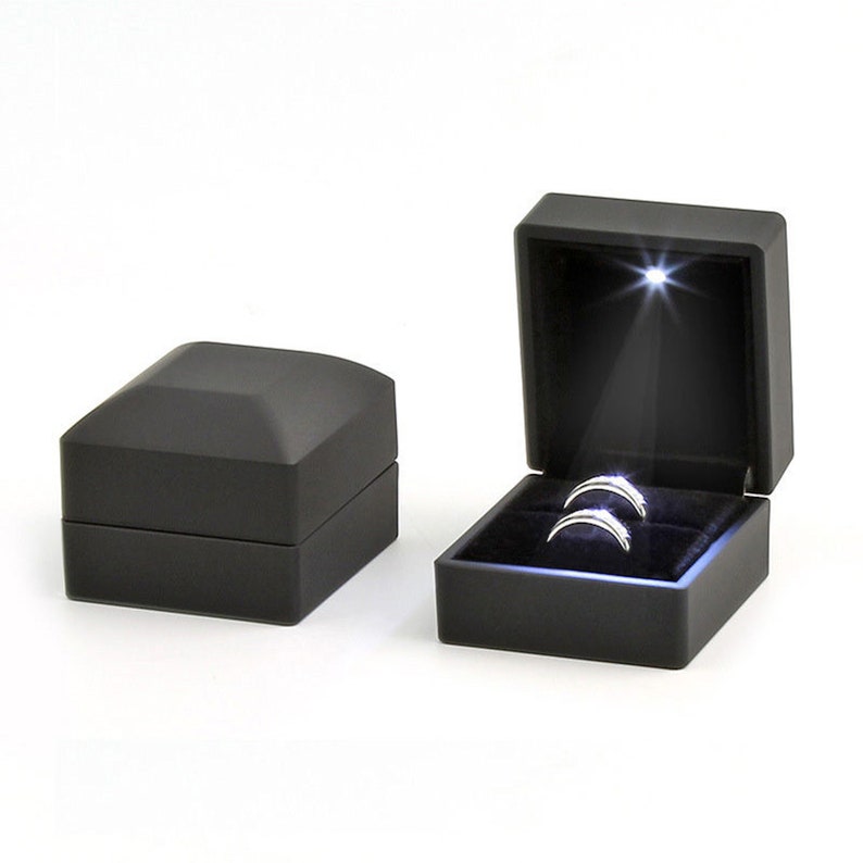 May include: Two silver rings displayed in an open black ring box with a spotlight shining on them. The box is velvet-lined and has a hinged lid. A closed black box is next to the open box. The boxes are on a white background.