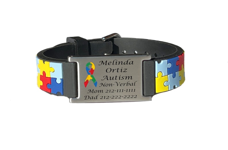 May include: Black and gray identification bracelet with a puzzle piece design in primary colors. The metal plate is engraved with the name "Melinda Ortiz", the word "Autism", and contact information. A colorful ribbon symbol is also present.