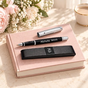 Puede incluir: Un bolígrafo negro con detalles plateados y el nombre Michelle Smith grabado en el cuerpo, acompañado de una tapa plateada a juego y un estuche rectangular negro con el monograma MS, todo sobre un cuaderno de color rosa.