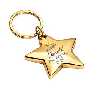 May include: A gold-colored star-shaped keychain with a graduation cap and scroll design. The keychain has the name "Donald" and the text "Proud Of You 2026" engraved on it. The keychain is attached to a gold-colored ring.