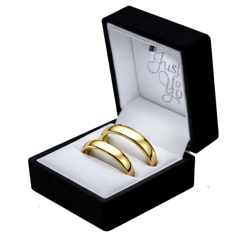 Puede incluir: Dos alianzas de boda doradas presentadas en un joyero negro abierto con interior blanco. La caja tiene el texto "Just For You" en escritura plateada. Los anillos son lisos y curvados, reflejando la luz.