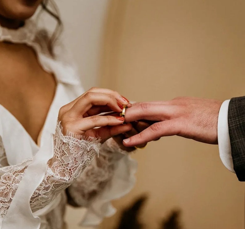 Puede incluir: Primer plano de una ceremonia de boda. Una mano coloca una alianza de oro en el dedo de otra persona. La mano lleva una manga de encaje blanco. La otra persona lleva un traje gris.