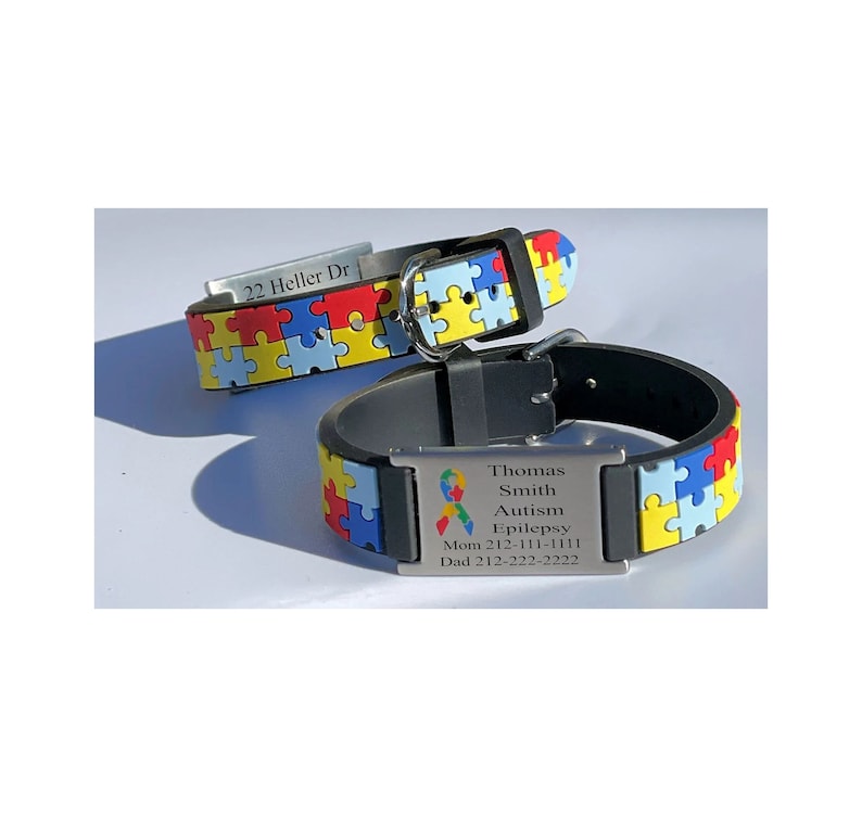 May include: Two black and puzzle-patterned bracelets. One bracelet has the text "22 Heller Dr" on a silver plate. The other bracelet has the text "Thomas Smith Autism Epilepsy Mom 212-111-1111 Dad 212-222-2222" on a silver plate.