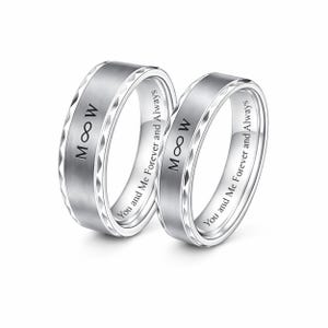 May include: Two silver wedding bands with the inscription "M &infin; W" and "You and Me Forever and Always". The rings have a brushed center and a geometric pattern on the edges.