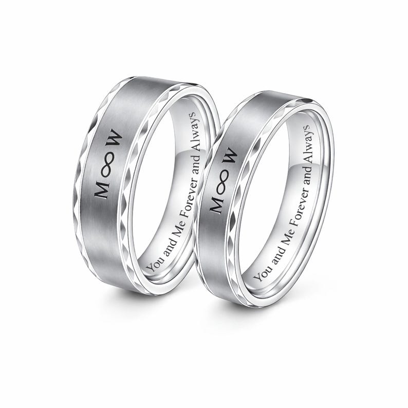 May include: Two silver wedding bands with the inscription "M &infin; W" and "You and Me Forever and Always". The rings have a brushed center and a geometric pattern on the edges.