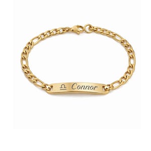 May include: Gold-colored bracelet with a Figaro chain design. The bracelet features a rectangular plate engraved with the name "Connor" and a Libra zodiac symbol. The bracelet is made of a shiny, gold-toned metal.