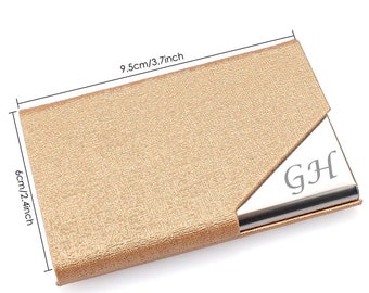 May include: A gold metallic card holder with a silver metal insert. The insert has the initials "GH" engraved on it. The card holder measures 9.5 cm by 6 cm.