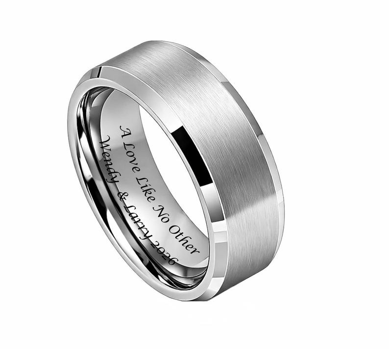 May include: A silver-colored ring with a brushed metal finish. The ring has a flat top and beveled edges. Engraved on the inside is the text "A Love Like No Other Wendy & Larry 2026".