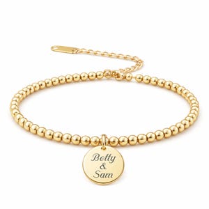 May include: Gold-colored beaded bracelet with a round charm. The charm is engraved with the names "Betty & Sam". The bracelet is made of small, round beads and has an adjustable chain closure. A simple and elegant piece of jewelry.