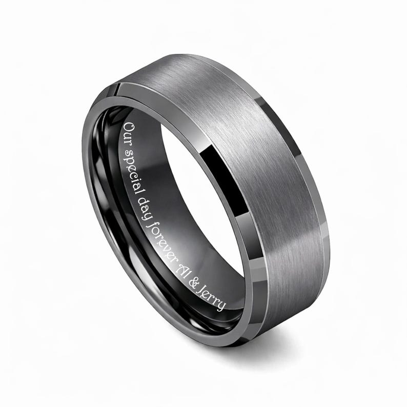 May include: A gray tungsten ring with a brushed finish and beveled edges. The inner band is black with the inscription "Our special day forever Al & Jerry". The ring has a modern, minimalist design.