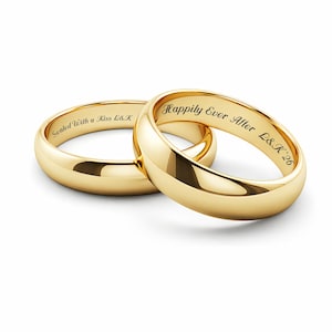 Puede incluir: Dos anillos de boda dorados con inscripciones. Uno dice "Sealed With a Kiss L&K", y el otro "Happily Ever After L&K '26". Los anillos son lisos y brillantes, reflejando la luz.