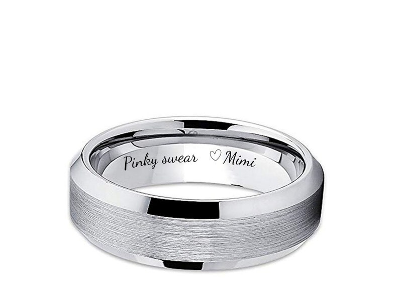 May include: A silver-colored ring with a brushed center band and polished edges. The inside of the ring is engraved with the words "Pinky swear" and "Mimi" with a heart symbol. The ring is a classic style.