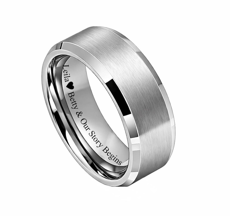 May include: A silver-colored ring with a brushed metal finish. The ring has a flat top and beveled edges. Engraved on the inside is the text "Leila ❤ Betty & Our Story Begins".